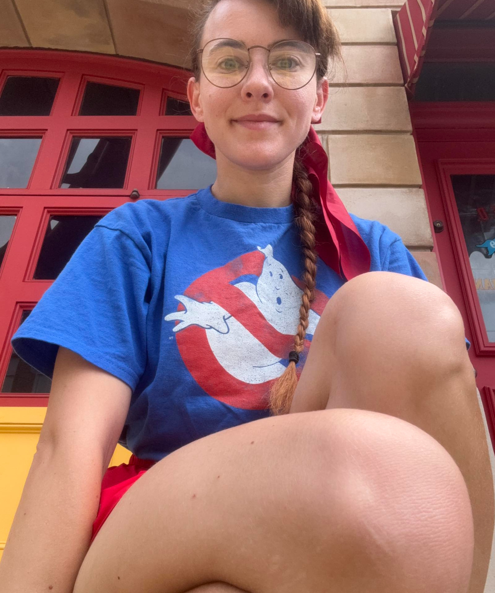 Ali, a female-presenting small human with auburn hair in a single braid with a red bow draped on their left shoulder. They are squatting in front of a firehouse door and are wearing a blue shirt with the "No Ghosts" logo from Ghostbusters on it and red sho
