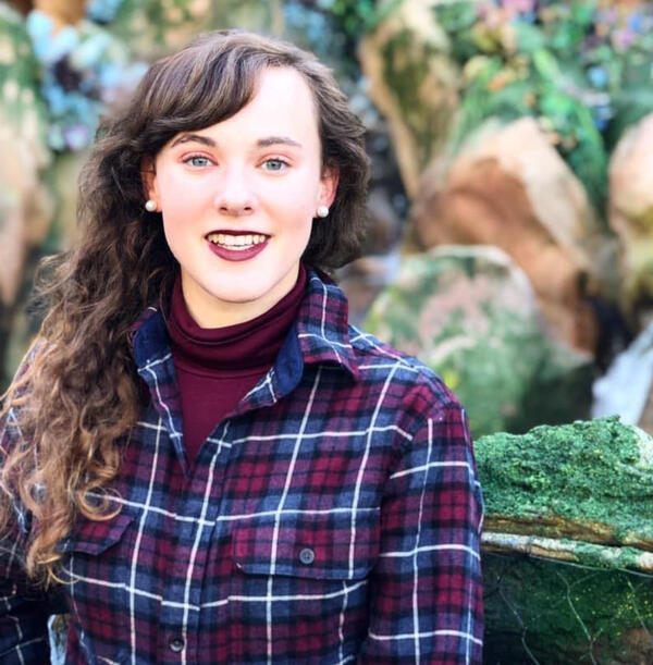 Ali, a female-presenting small human with long, curly light brown hair stands in front of mossy stones. She is wearing a blue and burgundy plaid shirt with a burgundy turtleneck underneath. She is smiling and has burgundy lipstick on. She has pearl earring