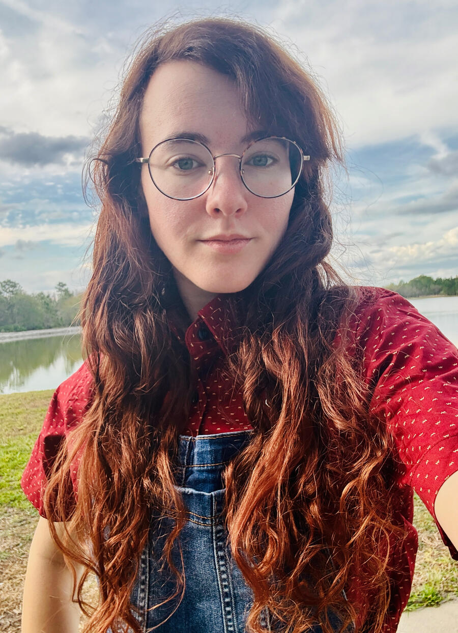 Ali, a female-presenting small human with long, curly auburn hair that is draped over her shoulders. She is wearing a red polka dot button up and jean overalls and a pair of oval glasses. She is slightly smiling (no teeth).
