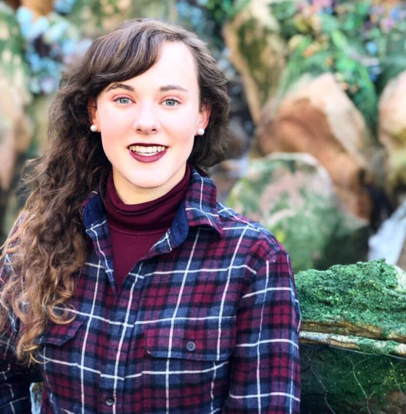 Ali, a female-presenting small human with long, curly light brown hair stands in front of mossy stones. She is wearing a blue and burgundy plaid shirt with a burgundy turtleneck underneath. She is smiling and has burgundy lipstick on. She has pearl earring