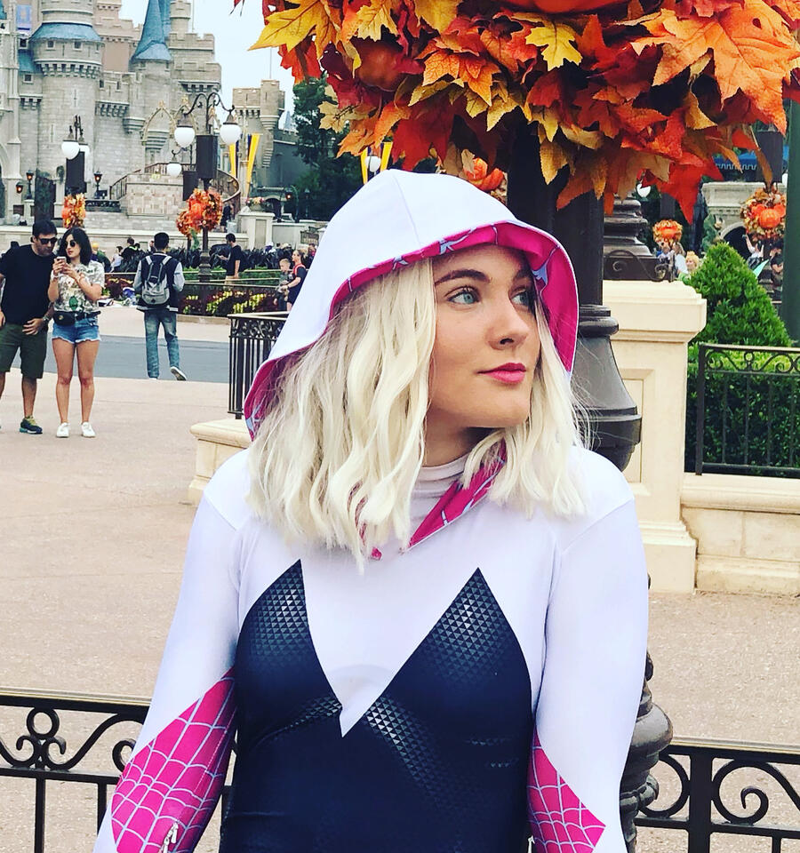 Ali, a female-presenting small human with a shoulder-length platinum blonde wig is in front of a Mickey pumpkin and Cinderella Castle. She is wearing a Spider-Gwen suit and subtle makeup, and is looking off in the distance with a nuetral expression on her