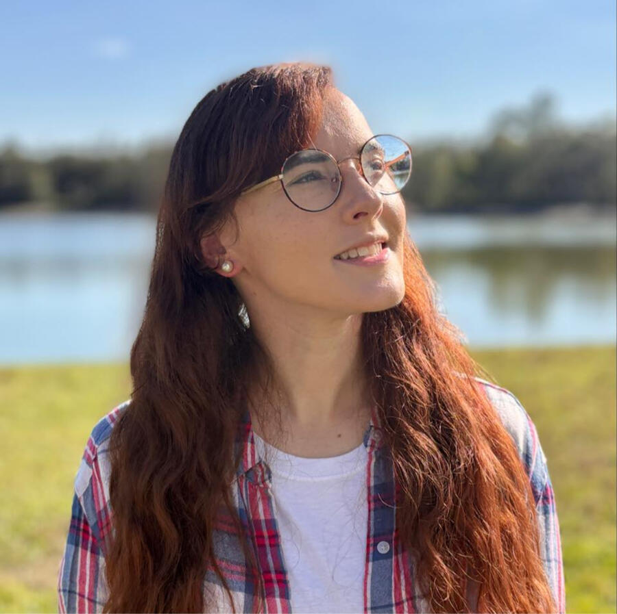 Ali, a female-presenting small human with curly, long brown hair is in front of mossy stones. She is wearing a blue and burgundy plaid shirt with a burgundy turtleneck underneath. She is smiling and has burgundy lipstick on as well as pearl earrings.